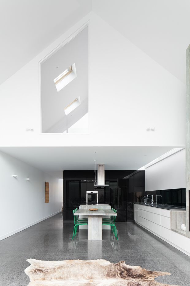 Bright contemporary open-plan kitchen and dining space with a dramatic double-height void above. The room has glossy black full-height cabinetry, a long white kitchen run with integrated appliances, a central stone island with green dining chairs, polished dark flooring, white walls, and high-level skylights. A partial stair or upper-level opening is visible above the kitchen, emphasizing the vertical volume.