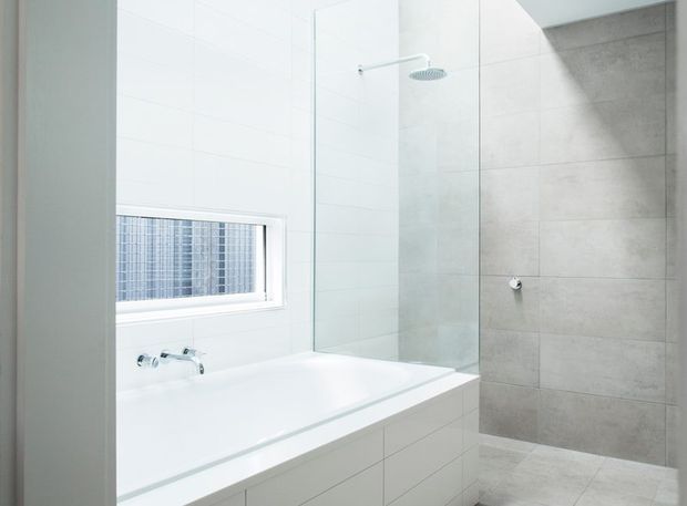 Bright, minimalist contemporary bathroom featuring a white tiled wall, built-in white bathtub with wall-mounted chrome fixtures, frameless glass shower screen, and a separate shower zone finished in large grey stone-look tiles. A horizontal frosted or textured window provides natural light.