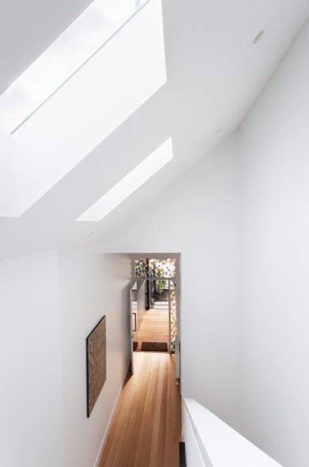 A bright, minimalist interior hallway or landing with white walls and ceilings, warm timber flooring, a large skylight and a long linear rooflight illuminating the passage. A framed artwork is mounted on the wall, and the corridor opens toward a timber outdoor deck or glazed area in the distance. The space has a strong sense of vertical openness and clean architectural geometry.