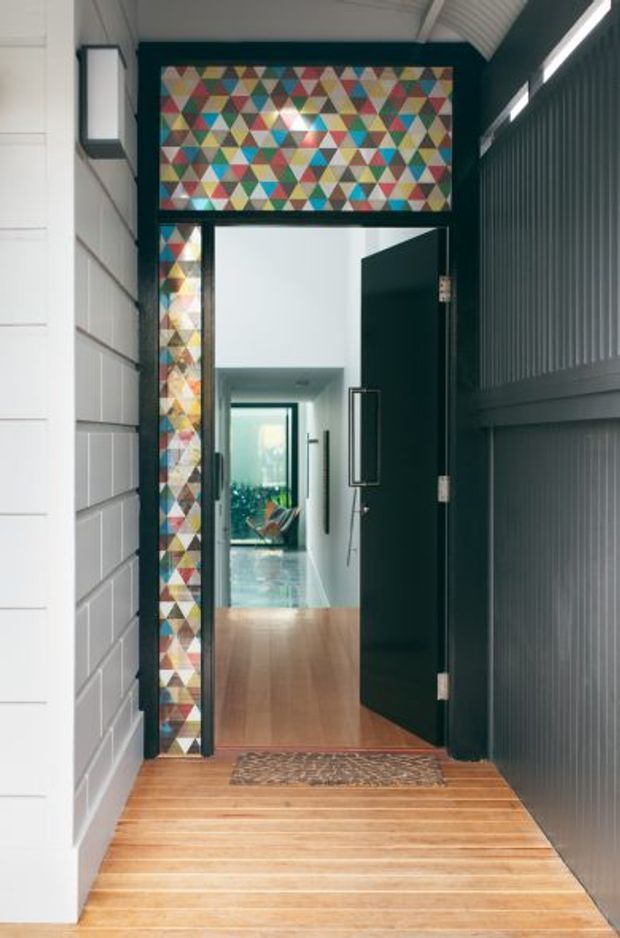 A contemporary residential entry viewed from an exterior threshold, with a warm timber-plank porch leading to a tall dark entry door. The doorway is framed by a colourful geometric stained-glass transom and sidelight, contrasting with white horizontal cladding on the left and dark vertically ribbed wall panelling on the right. A timber-floored hallway and glazed interior space are visible beyond.