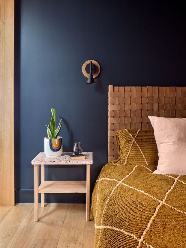 A warmly styled bedroom vignette featuring a woven leather or timber-look bedhead, mustard ochre textured bedding with cream geometric lines, and a pale blush cushion. A small timber bedside table has a terrazzo-style top and open lower shelf, holding a potted plant and glass. The room has a deep navy painted wall, light timber flooring and matching timber wall trim, with a compact black and brass wall sconce. The composition is intimate, tactile and strongly colour-blocked.