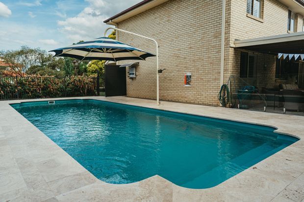 Exterior view of a residential swimming pool beside a light brick two-storey house. The pool is surrounded by pale paving, with a large striped umbrella, poolside seating, a covered patio, and garden screening visible in the background.