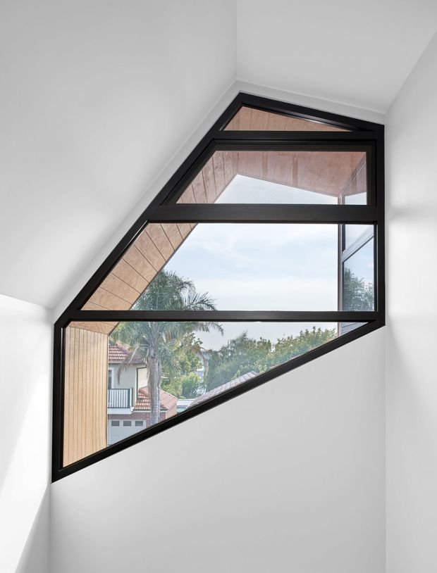 A bright, minimalist interior stairwell or landing dominated by a dramatic custom geometric window. The angular black-framed glazing follows the sloping ceiling and stair geometry, bringing views of neighbouring homes and trees into an otherwise white space. Warm timber soffit/cladding is visible through the window.