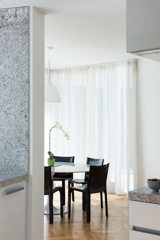 Bright, refined dining area viewed from an adjacent kitchen. A round glass dining table with dark upholstered dining chairs sits in front of full-height sheer curtains, with a white pendant light overhead. The surrounding kitchen includes pale cabinetry and prominent speckled stone benchtops, while the dining area has a light timber parquet floor and white walls.