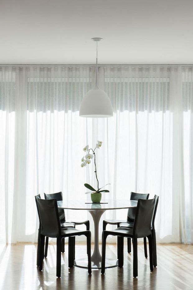 Bright, minimalist dining area with a round glass dining table, sculptural pedestal base, four black dining chairs, and a potted orchid centerpiece. Floor-to-ceiling sheer curtains provide soft natural light, while a simple white pendant hangs above. The room has pale timber flooring and a restrained monochromatic palette.