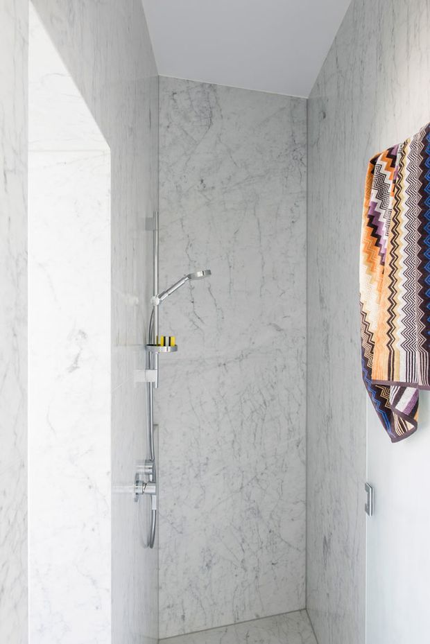 A narrow contemporary shower enclosure lined floor-to-ceiling in pale grey-white marble with pronounced natural veining. Chrome shower hardware, a handheld shower rail, recessed shelf, and a colourful geometric patterned towel are visible. The space is bright, minimal, and highly refined.