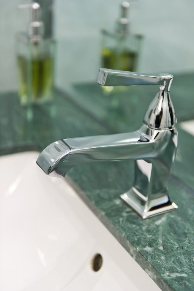 Close-up product-style photograph of a polished chrome bathroom basin tap mounted on a dark green veined stone countertop beside a white ceramic sink. Matching liquid soap dispensers and a reflective backsplash are visible in the background.
