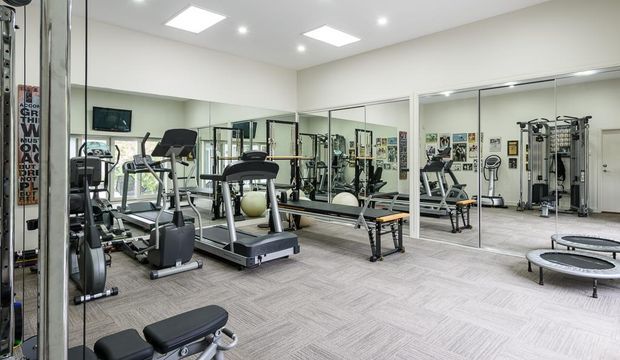 A bright, well-equipped home gym with multiple treadmills, elliptical machines, stationary equipment, a cable machine, weight benches and mirrored walls. The room has a light neutral palette, recessed and square-panel ceiling lighting, and grey patterned carpet flooring.