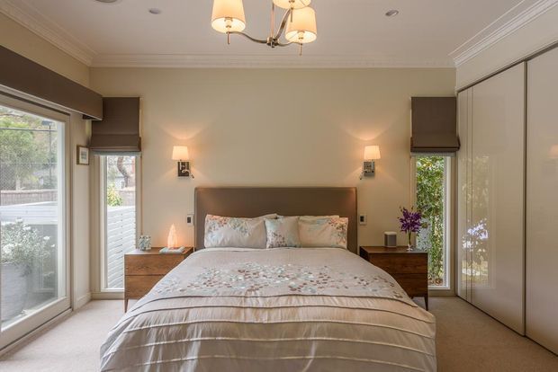 A warm, refined bedroom with a centered upholstered bed, layered cream and blush bedding, matching timber bedside tables, and wall-mounted bedside lamps. Full-height glazing on both sides provides garden views, while built-in wardrobes occupy the right wall. The room has a decorative ceiling cornice and a traditional chandelier.