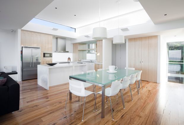 Bright open-plan kitchen and dining area with white stone island cabinetry, integrated stainless-steel appliances, pale timber joinery, a glass dining table with white chairs, and warm timber flooring. A large skylight or clerestory glazing and suspended white pendant lights create an airy contemporary interior.
