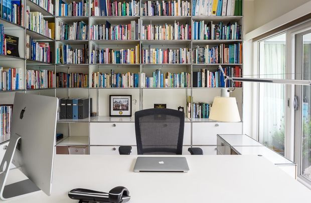Bright, modern home office with a large white desk, desktop computer, laptop, ergonomic mesh chair, and extensive wall-to-wall built-in bookshelves filled with books. White built-in cabinetry and shelving line the back wall, while large glazed doors or windows provide abundant natural light on the right.