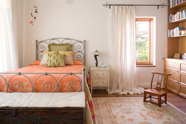 A warm, traditionally styled bedroom with an ornate wrought-iron bed, coral-orange patterned bedding, layered cushions, a small bedside table and lamp, a wooden chest at the foot of the bed, and a built-in wooden bookcase and dresser. Soft cream curtains frame a timber-trimmed window, while a vintage wooden chair and patterned rug add character.