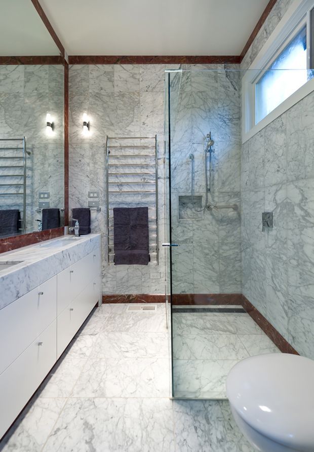 Luxurious contemporary bathroom clad extensively in veined white marble, with matching marble floor and walls, dark reddish-brown marble trim, a frameless glass walk-in shower, wall-mounted shower fittings, floating white vanity cabinetry, mirrored wall, heated towel rail, and a high horizontal window.