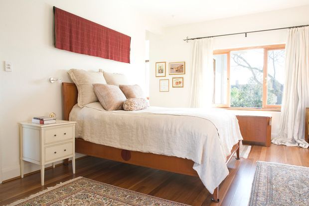 Bright, warm bedroom with a timber bed, layered neutral linen, white bedside chest, hardwood flooring, patterned rugs, and a large timber-framed window dressed with sheer curtains. A red textured wall artwork provides a bold focal accent, while a low timber cabinet sits beneath the window.