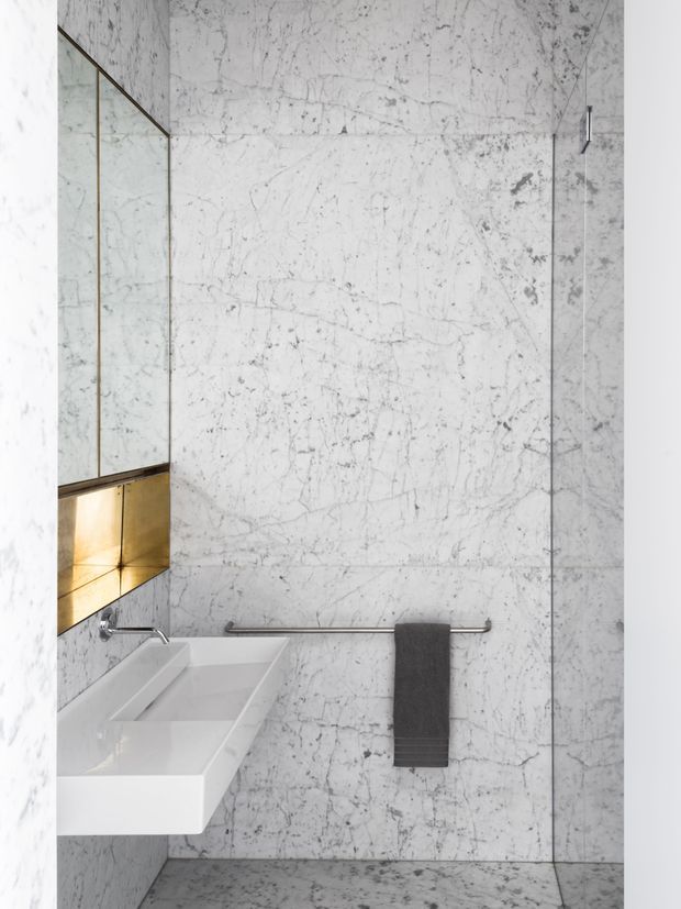A refined contemporary bathroom featuring full-height white marble wall and floor surfaces with prominent grey veining. A frameless glass shower enclosure occupies the right side, while a white rectangular wall-mounted basin sits beneath a large mirror on the left. A recessed metallic gold-toned niche adds a warm accent, complemented by chrome tapware and towel rail and a charcoal towel.