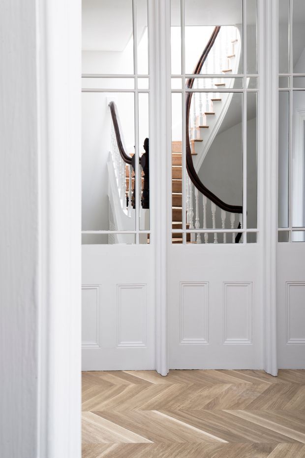 Bright, refined interior view of a hallway or stair landing seen through white glazed-panel internal doors. The doors feature multiple divided glass panes above recessed lower panels, opening onto a traditional staircase with white turned balusters and a dark timber curved handrail. The foreground has a light natural timber herringbone parquet floor and white walls and trim.
