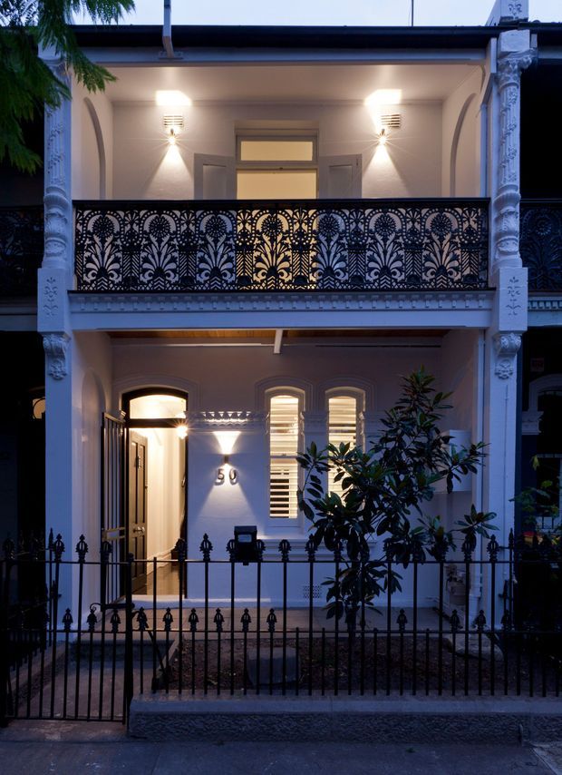 A dusk exterior view of a narrow two-storey terrace-style residence. The facade features ornate white-painted columns and moulded detailing, arched openings, shuttered windows, a recessed entry porch, and a highly decorative black metal balcony balustrade with floral motifs. A front garden and low wrought-iron fence sit in front of the illuminated home.