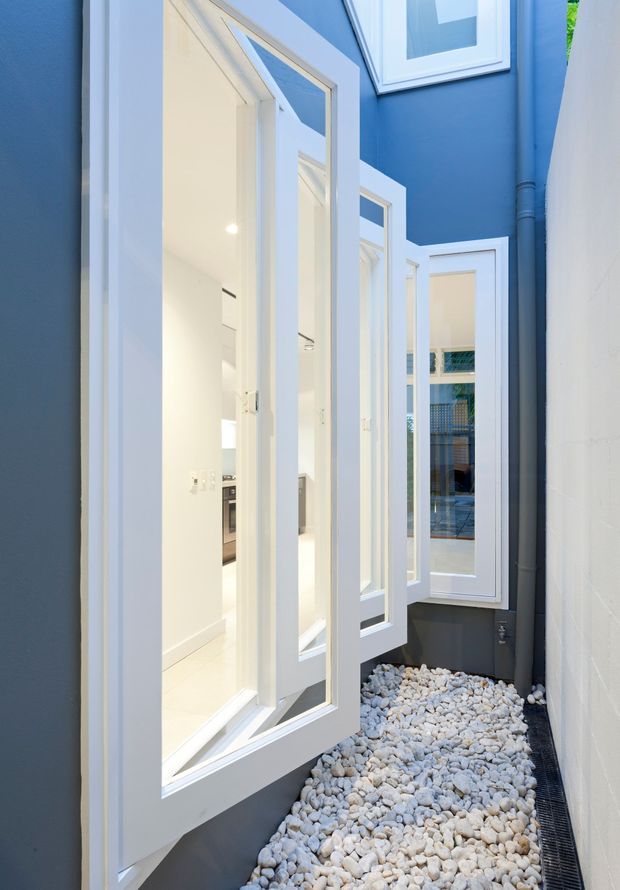 A narrow, bright lightwell or courtyard passage between blue-painted walls, featuring multiple tall white-framed glazed windows and doors opening into the adjoining interior. The floor is covered with white decorative stones and a linear drainage channel runs along the edge. The image highlights the crisp contrast between deep blue walls, white joinery, and pale gravel.