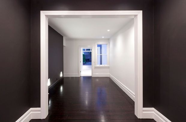 A narrow, empty interior passage or small room viewed through a wide white-trimmed opening. The space has glossy dark timber flooring, white walls and ceiling, dark charcoal wall panels or paint at the sides, layered decorative skirting and architraves, recessed ceiling lights, and a small window and glazed door at the far end. The overall presentation is clean, high-contrast, and refined.