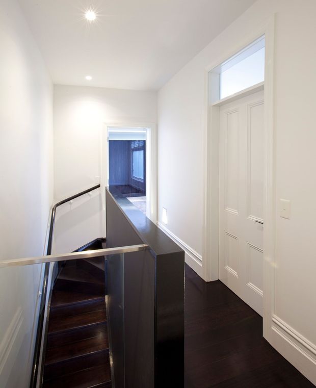 Bright, narrow upper-level landing beside a modern stairwell. The space has white walls and ceiling, recessed and spotlight lighting, dark timber flooring and stair treads, white panelled internal doors with a high transom window, and a glass stair balustrade with dark metal or timber handrails. An adjoining room is visible through the doorway at the end of the landing.