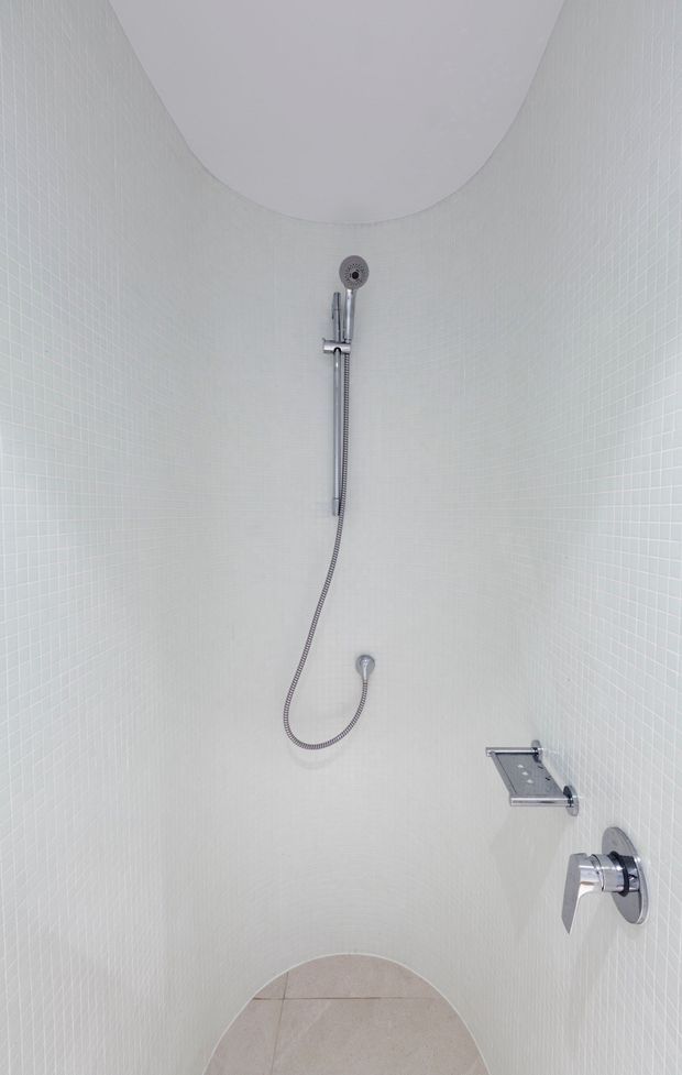 A compact minimalist shower enclosure with curved walls and a rounded ceiling detail, fully lined in small white mosaic tiles. Chrome shower fixtures, handheld shower hose, wall-mounted control, and a narrow glass shelf create a clean, bright bathroom interior.