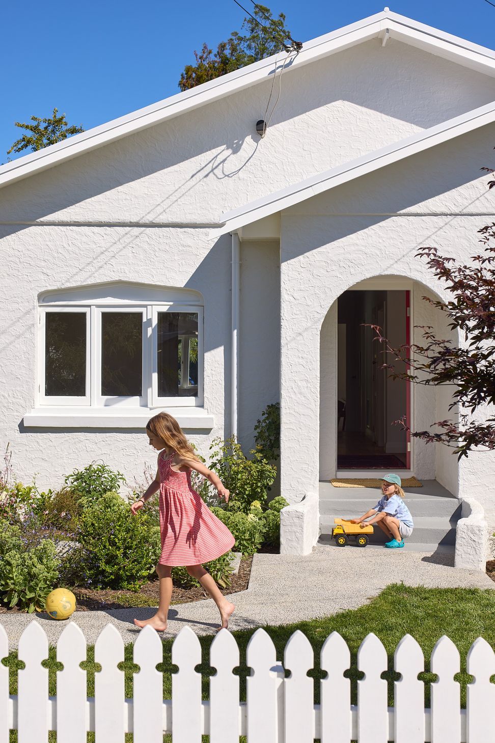 The restored bungalow provides a welcoming family setting, with children playing in the front garden and the distinctive red front door standing open. The landscaped garden reinforces the home's friendly neighbourhood character.