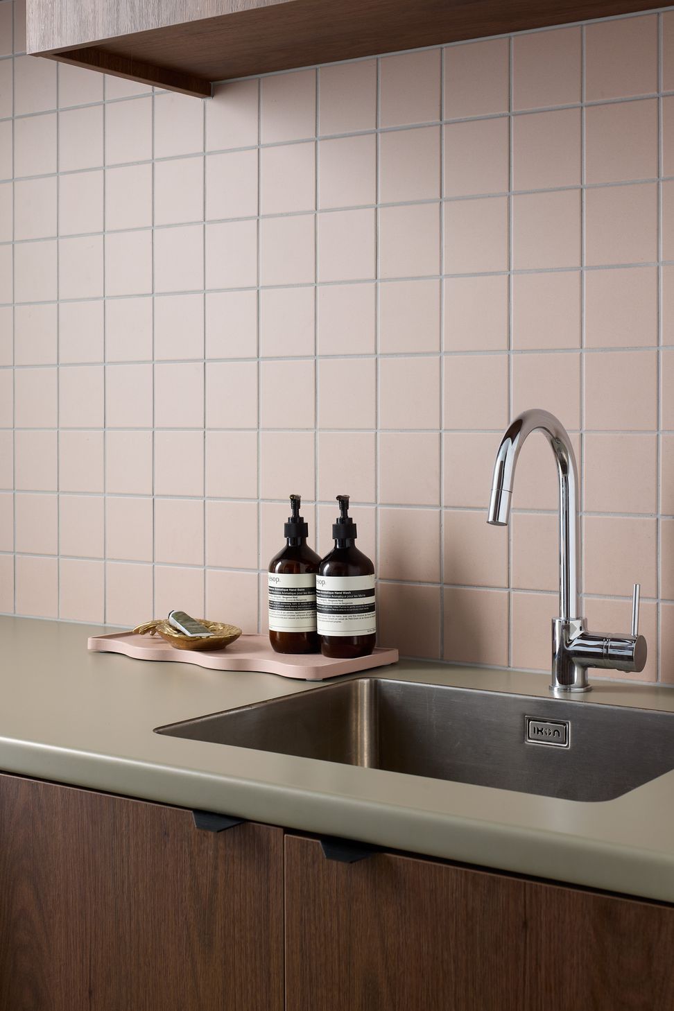 The laundry continues the home's playful material palette, combining a green benchtop with walnut cabinetry and soft pink wall tiles in a compact yet beautifully detailed workspace.