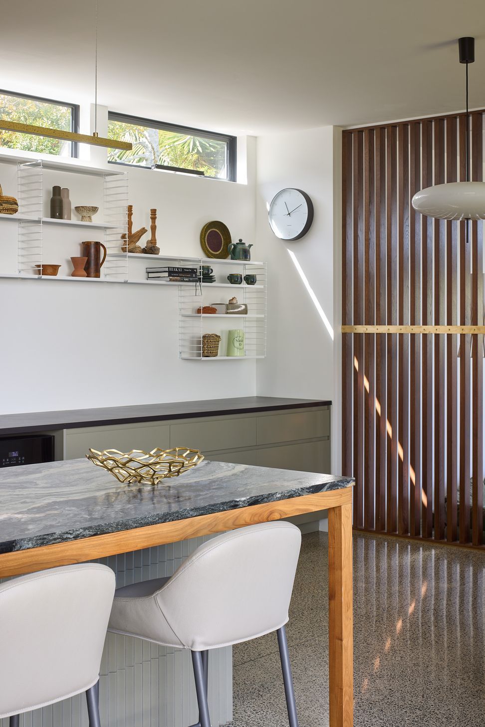 Standing behind the kitchen island, views extend towards the bespoke dining cabinetry with white metal shelving. The sculptural timber screen beyond creates depth and visual connection throughout the open-plan interior.