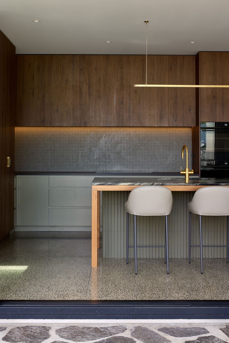 Viewed from the garden, the kitchen's warm walnut cabinetry, green joinery, brass hardware and expansive glazing create an inviting connection between indoors and out.