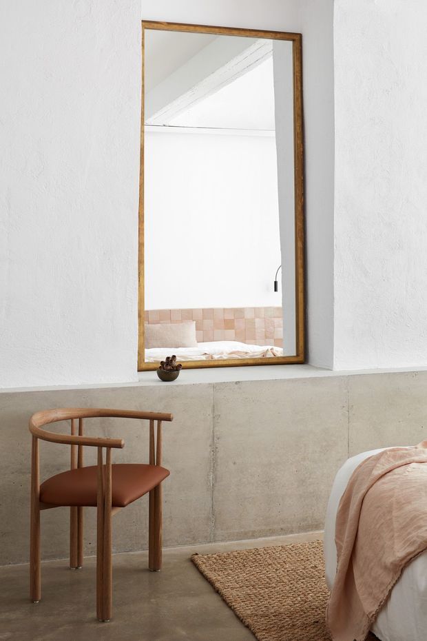 A softly lit bedroom corner with white textured plaster walls, a low raw concrete half-wall, and a polished concrete floor. A large rectangular mirror with a warm timber frame reflects the bed, white ceiling structure, and a small pendant light. In the foreground is a sculptural timber chair with a brown leather seat, beside a woven natural-fibre rug and the edge of a bed dressed in pale blush bedding. The restrained palette and natural materials create a calm, minimalist interior.