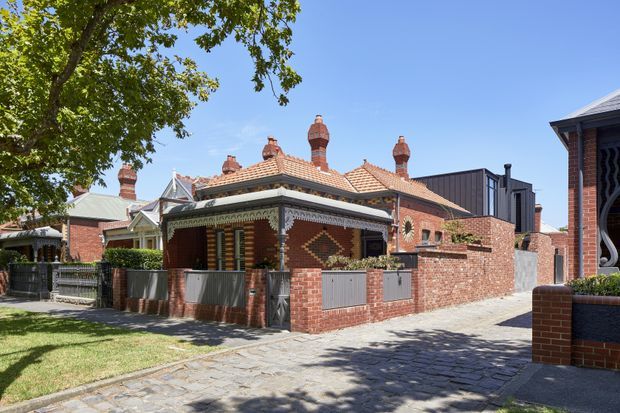 Street-level view of a historic red-brick terrace or cottage with a low tiled roof, multiple ornate brick chimneys, decorative verandah detailing, patterned brickwork, and a small front garden enclosed by brick and metal fencing. Adjacent heritage-style residences and a paved driveway are visible, with mature trees and clear daylight.