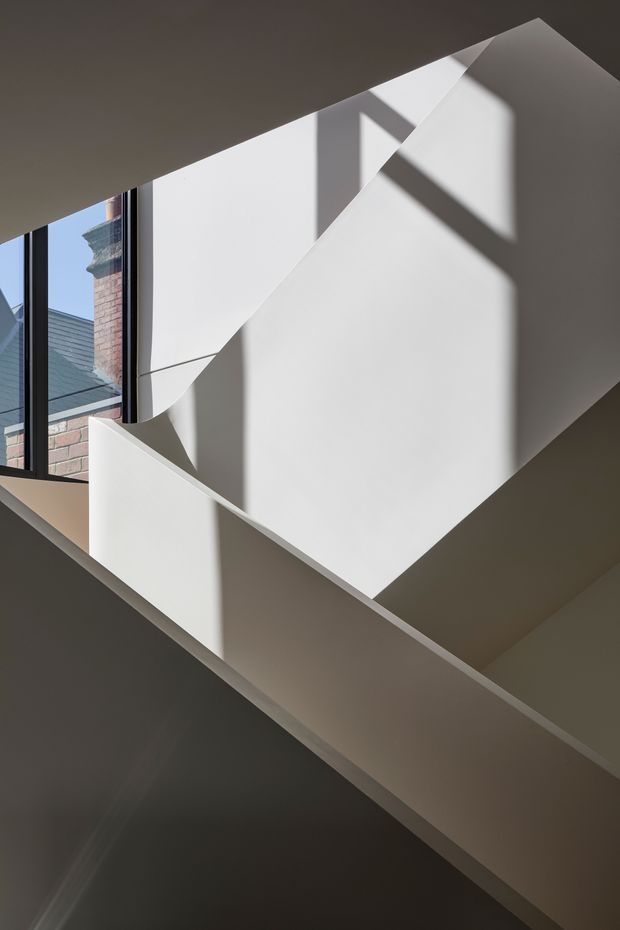 Abstract architectural view of a stairway or landing, featuring crisp white geometric planes, angular balustrades, strong sunlight, and dramatic cast shadows. A tall dark-framed window reveals neighbouring brick and slate-roofed buildings outside. The composition emphasizes modern spatial geometry and high contrast rather than furnishings.