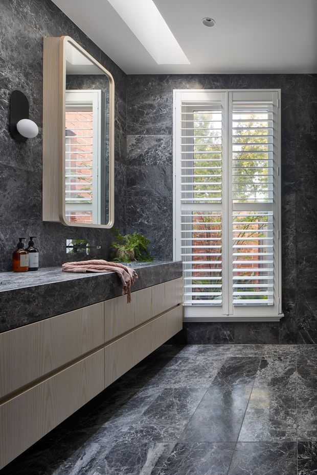 A refined contemporary bathroom featuring a long floating timber-toned vanity with integrated dark stone benchtop and backsplash, large-format dark veined stone wall and floor surfaces, a rounded rectangular illuminated mirror, and white plantation shutters over a tall window. A skylight introduces natural light, while black-and-white wall lighting and restrained accessories add a luxurious, moody character.