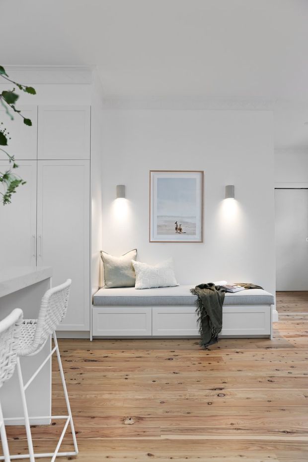 Bright, minimalist white interior featuring a built-in window-style bench seat with drawers and cushions, tall integrated cabinetry, a white kitchen or counter edge with woven bar stools, pale timber flooring, wall-mounted sconces, and framed coastal artwork. A leafy indoor plant and soft textiles add subtle warmth.