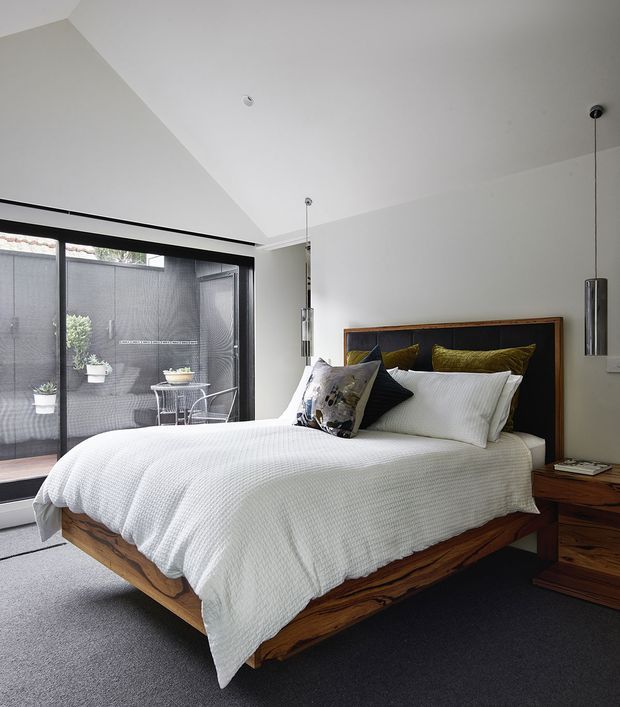 A bright contemporary bedroom with a large timber bed featuring a thick live-edge-style frame and matching timber bedside furniture. White textured bedding and pillows contrast with dark charcoal carpet and a warm wood headboard. Full-height black-framed glazing opens toward a small planted courtyard or balcony, while slim cylindrical pendant lights hang beside the bed. The room has a clean, calm, refined atmosphere with a sloped ceiling and abundant natural light.