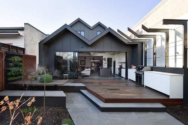 Contemporary residential rear facade with a distinctive multi-gabled dark-clad extension opening onto a broad timber deck. The outdoor entertaining area includes a built-in white outdoor kitchen with barbecue, while large glazed doors connect to a bright open-plan interior. Layered paved steps, raised garden beds, planting, and neighbouring masonry and timber elements frame the view.