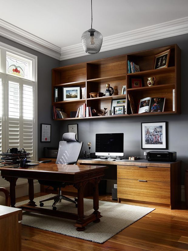 A refined home office with dark charcoal-grey walls, ornate white crown moulding, a tall shuttered window with decorative stained glass, and warm timber flooring. A large traditional carved timber desk sits in front of a contemporary office chair, while a wall-length custom timber shelving and cabinetry unit displays books, artwork, and decorative objects. The built-in desk area includes a computer, printer, and drawers, with a neutral textured rug beneath the workspace. The room combines traditional architectural detailing with warm, tailored contemporary joinery.