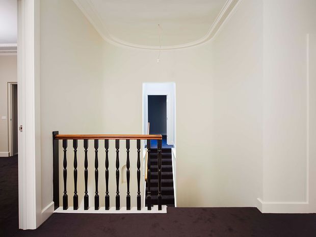 A bright traditional stair landing with ornate white wall mouldings and ceiling cornice, dark carpeted flooring, and a dark timber staircase balustrade with turned spindles and a polished wood handrail. A small stairwell opening and adjacent doorway are visible. The space is largely empty and refined, with strong period detailing.