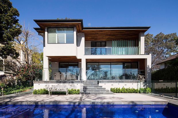 Contemporary two-storey residence with crisp white rendered walls, extensive glazing, glass balustrades, and broad flat roof planes. Warm timber soffits and screening add contrast. The home opens onto a paved terrace and a vivid blue swimming pool, with established trees and planting surrounding the site.