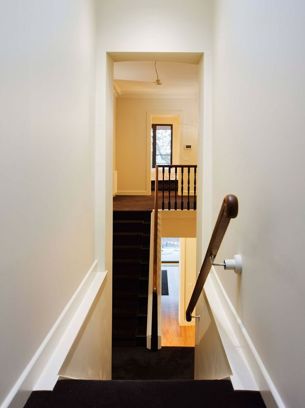 Interior view looking down a narrow carpeted stairway toward a lower-level landing and an adjacent staircase. The walls and trim are painted light neutral tones, with dark carpeted steps, a timber handrail, and black turned balusters. A window and polished timber flooring are visible beyond the stairwell.