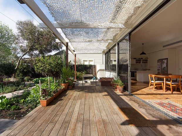 A sunlit timber deck and covered outdoor entertaining area beside an open-plan kitchen and dining room. The deck is framed by a lightweight pergola with a patterned shade canopy, screened glazing, potted plants, and a garden edge. Inside, timber dining furniture, white cabinetry, a tiled floor, and a pendant light are visible.