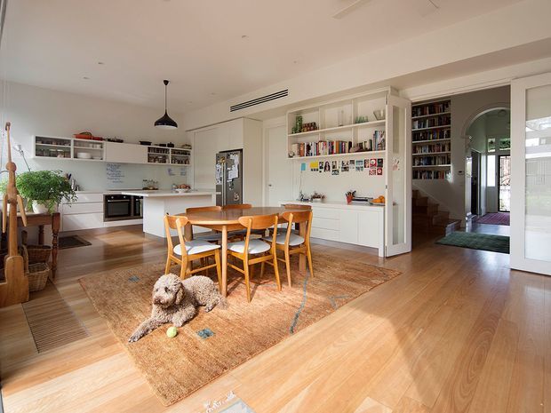 Bright open-plan kitchen and dining area with white built-in cabinetry, open shelving, a central island, timber dining table and chairs, and warm timber flooring. A living or entry area with an arched opening and staircase is visible beyond the joinery. The room includes a large natural-fibre rug, pendant light, indoor plants, and a dog.