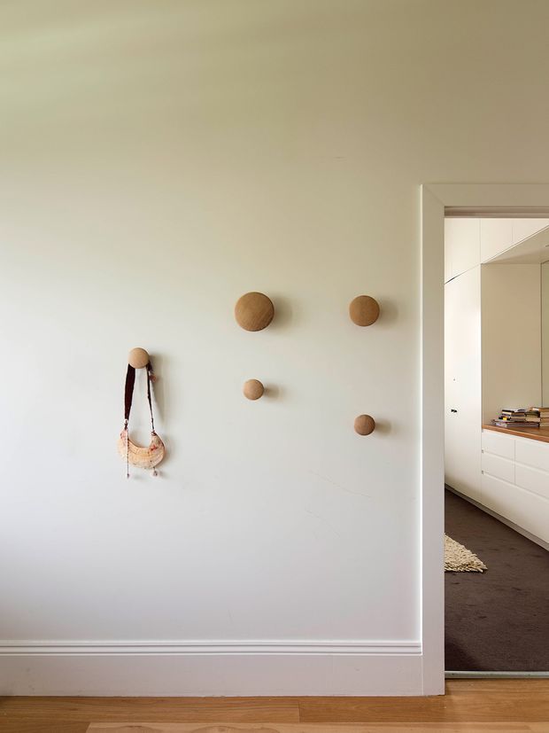 A minimalist interior view showing a pale painted wall with decorative round timber wall pegs and a hanging woven bag. White traditional-style skirting and trim frame the wall, while a timber floor runs across the foreground. An open doorway reveals a carpeted room with white built-in cabinetry and shelving beyond.