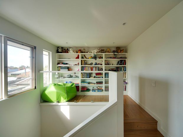A bright upper-level reading nook or study overlooking a stairwell, with extensive built-in white shelving filled with books, toys and objects. Large windows bring in natural light and offer suburban views, while a vivid lime-green beanbag provides seating. A timber stair and landing lead into the space, with smooth white walls and ceiling creating a clean, contemporary interior.
