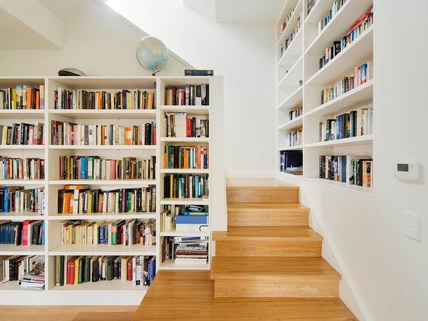 Bright contemporary stairway lined with extensive built-in white bookshelves filled with books. Warm timber stair treads and flooring contrast with the white walls and joinery, while a small globe sits atop the shelving. The stair rises toward a landing and is photographed from within the interior.