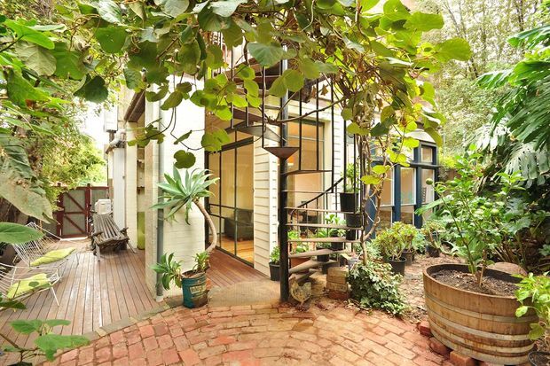 A lush, leafy courtyard and outdoor living area beside a compact multi-level building. Timber decking, a curved brick-paved path, dense tropical planting, a raised timber planter, and an open black metal spiral staircase frame pale weatherboard walls, large glazed doors, and dark-framed windows.