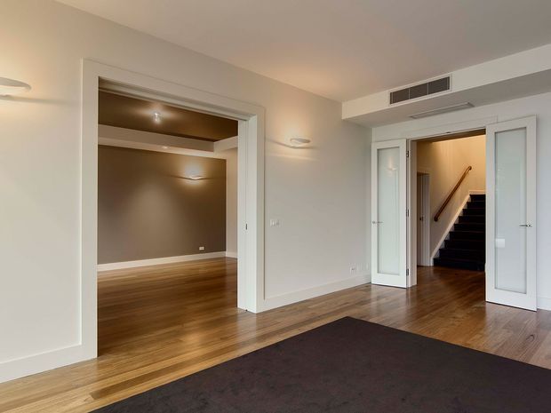 A bright, refined interior hallway or landing with warm timber flooring, pale painted walls, recessed and wall-mounted lighting, and white-trimmed openings. A dark carpeted area occupies the foreground. Double doors on the right open to a stairway, while a wide opening on the left leads to an additional polished timber-floored room with a darker feature wall.
