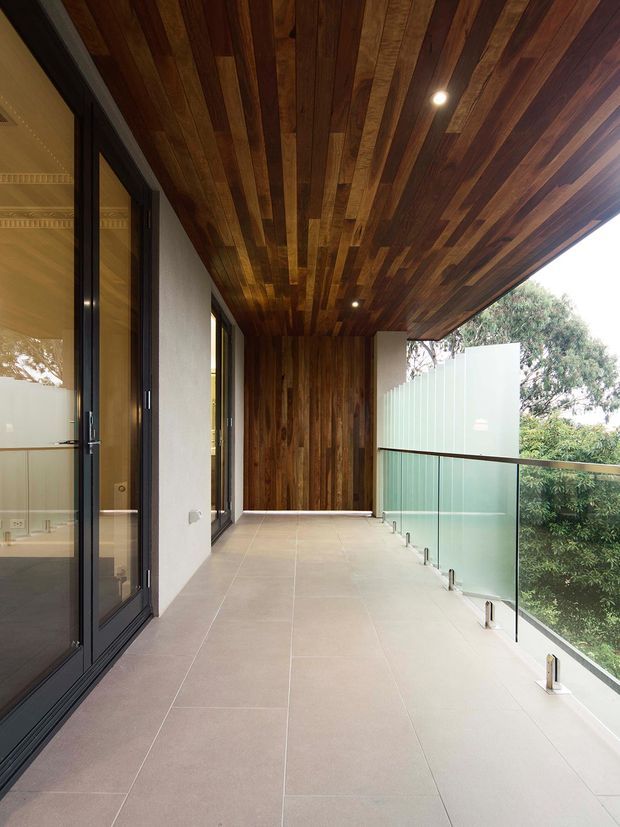 A covered outdoor balcony or terrace with large pale stone-look floor tiles, dark-framed glazed doors on one side, a full-height timber-lined ceiling extending to a timber feature wall, and frameless clear glass balustrades overlooking greenery. Recessed downlights provide illumination, creating a warm contemporary architectural detail.