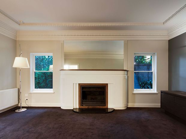 A formal, symmetrical living room with a central decorative fireplace and broad overmantel mirror, flanked by two sash windows. The room features cream painted walls, elaborate ceiling cornices and wall mouldings, dark brown carpet, a simple floor lamp, radiator, and dark timber storage furniture. The restrained palette and original detailing create a refined period interior.