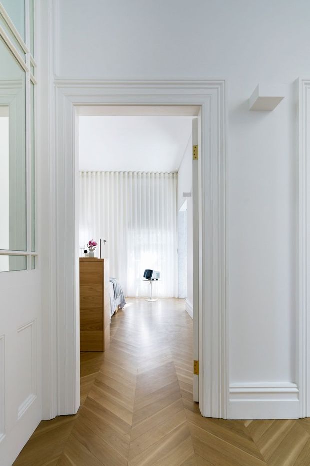 Bright, refined view through a heavily moulded white doorway into a bedroom. The room has pale herringbone timber flooring, sheer floor-to-ceiling curtains, a warm timber chest of drawers, and minimal furnishings. White wall panelling, decorative architraves, and a partially visible glazed door create a clean classic interior with abundant natural light.